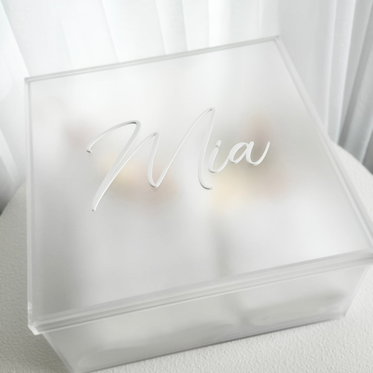 A frosted acrylic keepsake box with a custom name cutout on the lid, reading 'Mia', set against a soft white curtain background.
