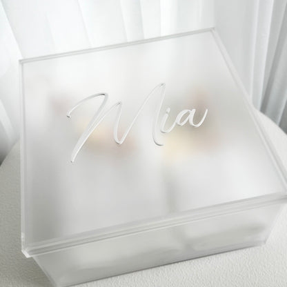 A frosted acrylic keepsake box with a custom name cutout on the lid, reading 'Mia', set against a soft white curtain background.